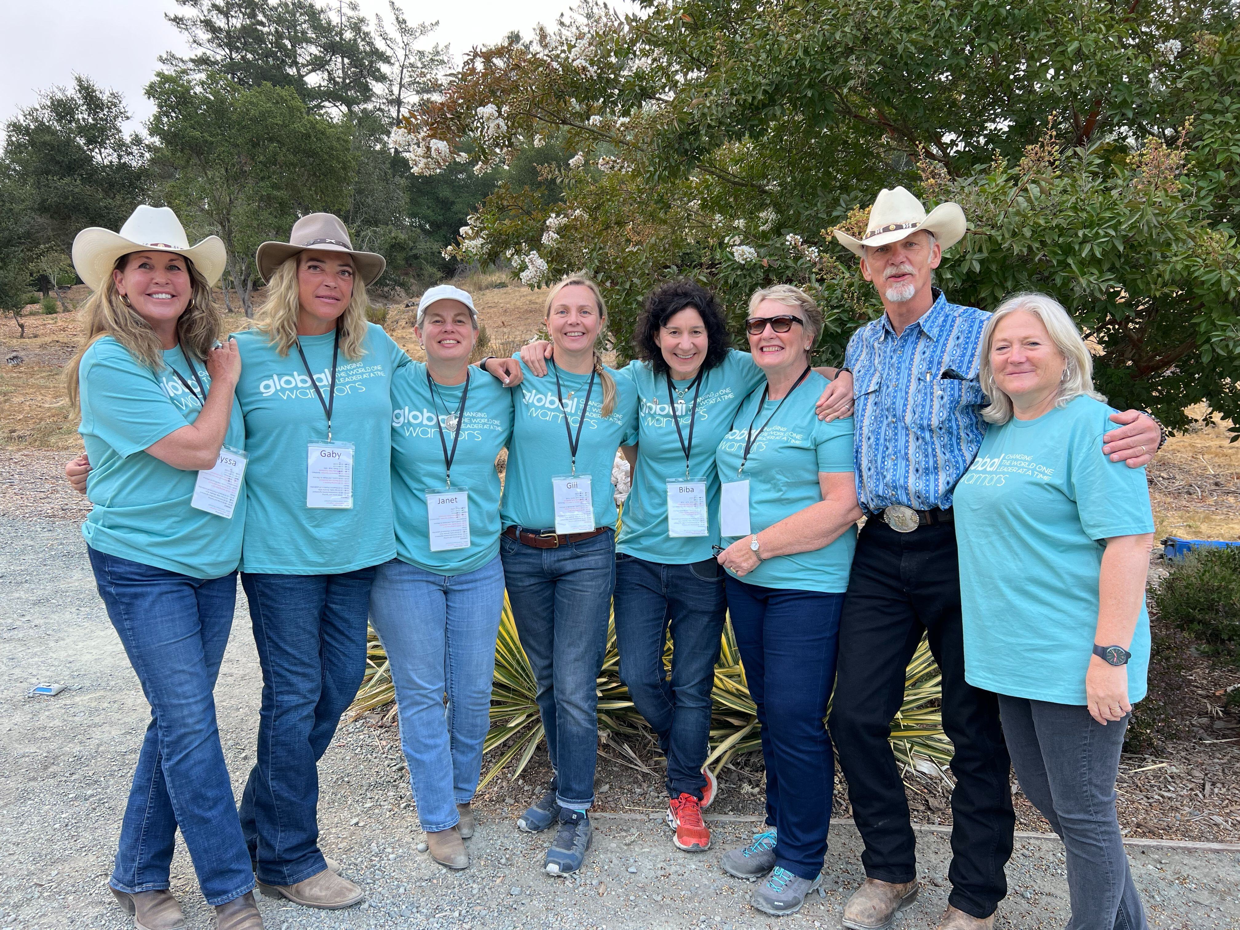 Eight members of the Global Warriors team standing together arms around each other, all dressed in blue t-shirts and jeans. One man and two women are also wearing cowboy hats.