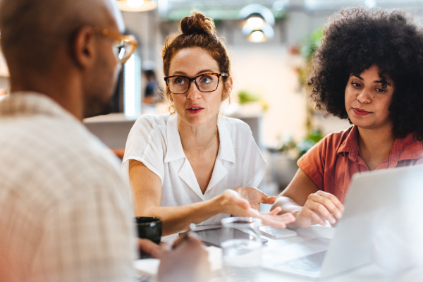 Three people at work in conversation. A man with his back to the camera and two women - one is looking at the man, the other at a laptop which is positioned between them.