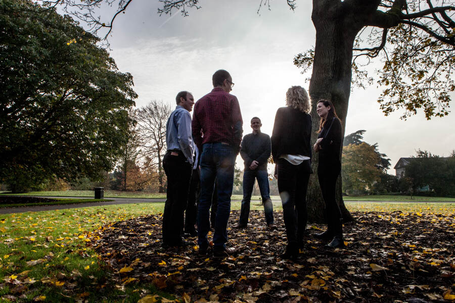 A group of people stood in a circle next to a tree on an autumn day. They are facing each other. 