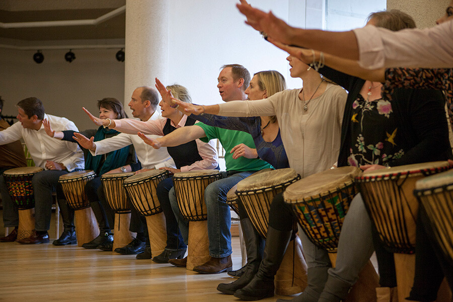 A group of people in a line sat playing the bongos. They have one arm raised in front of them. 