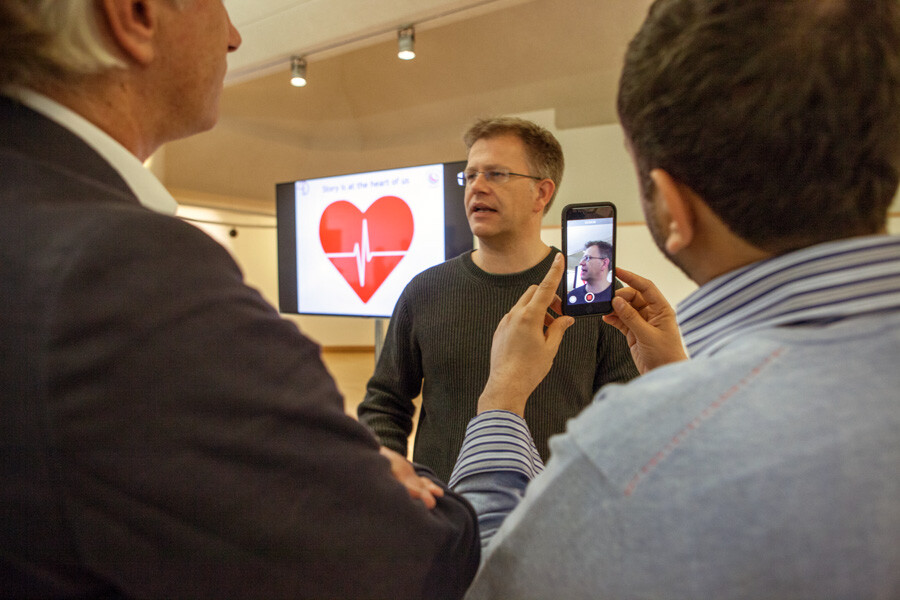 Two men with their backs to the camera, the third being filmed by a phone. Behind him a big screen with a large heart with a heartbeat through it. 