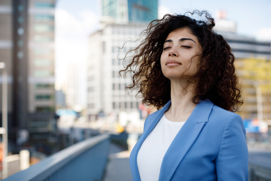 A woman at work standing with her eyes closed in a city. She appears to be taking a deep breath. 