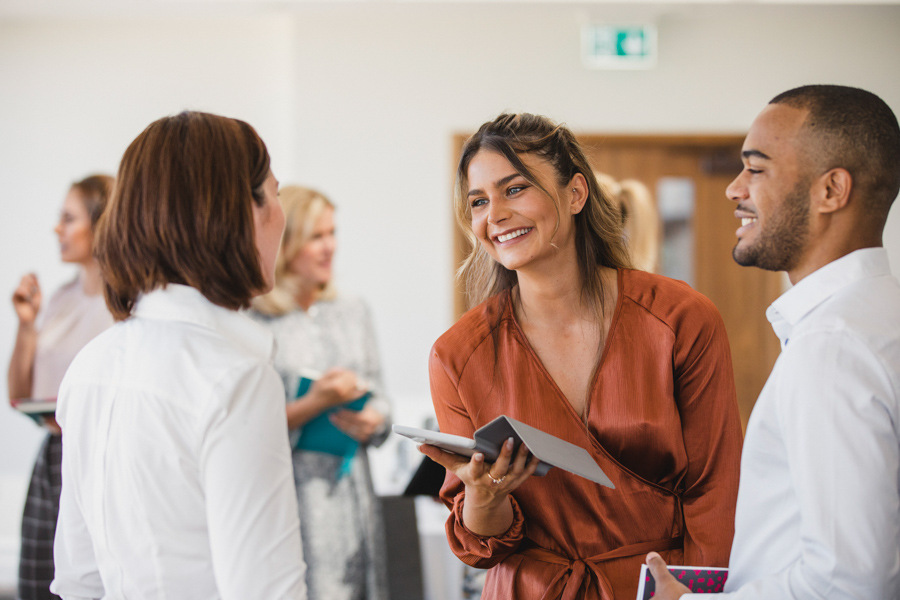 A woman at work standing and holding an ipad chatting to a man and woman with their backs to the camera. They are all smiling.