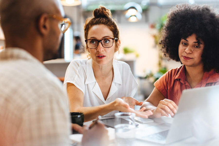 Three people working - A man and 2 women with a laptop in front of them. The man has his back to the camera. The first woman is looking at him and the second woman is looking at the computer screen. They appear to be discussing something. 