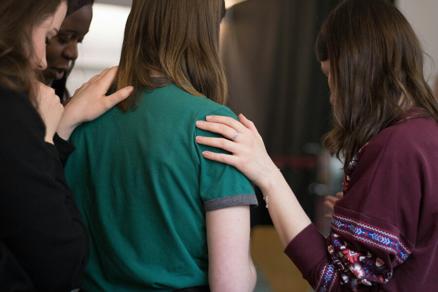 Three women all with their backs to the camera. Two women have a supporting hand on the should of the women in the centre of the image.