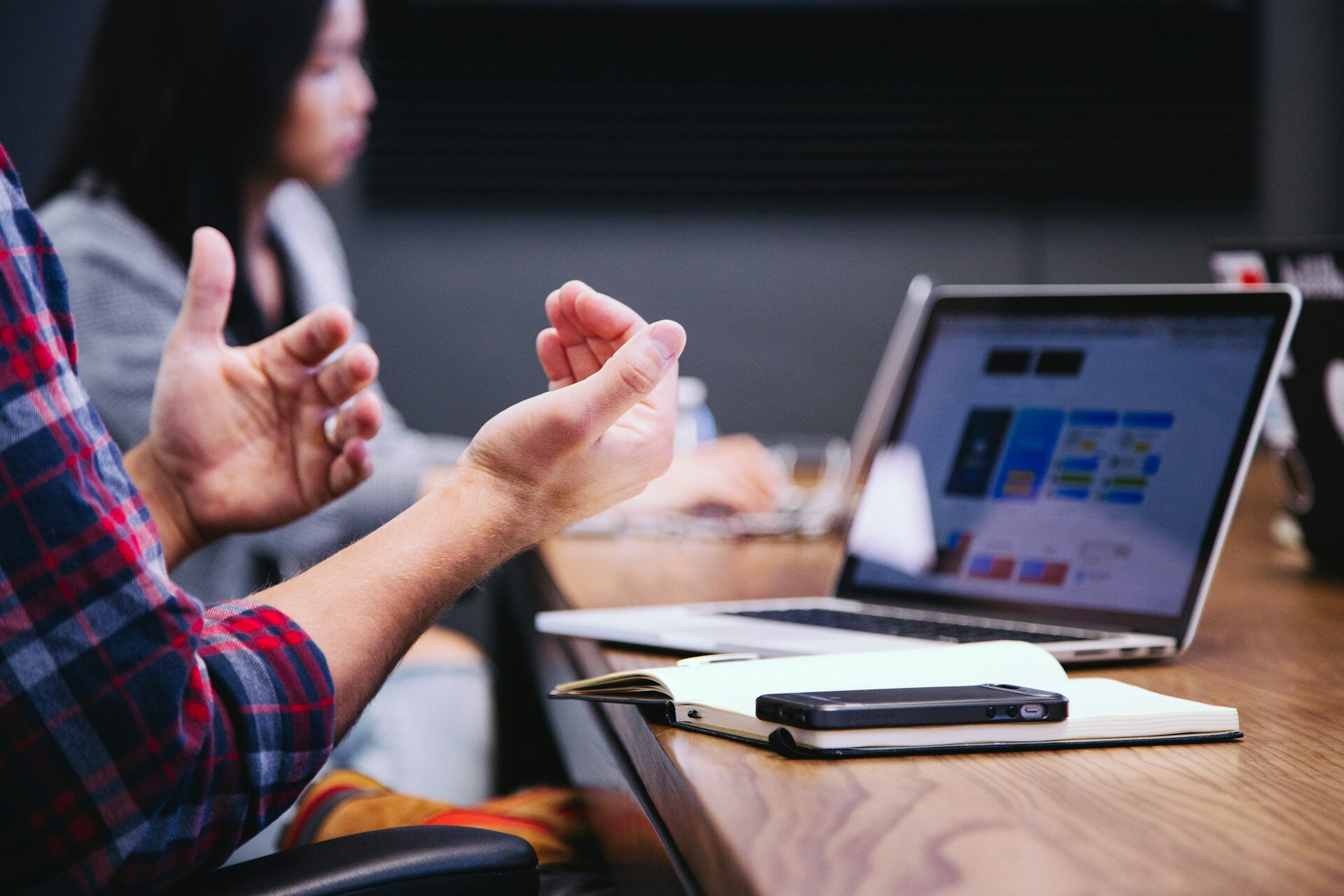 The image shows a persons hands being used to communicate, a desk, laptop and notepad. There is another person out of focus in the background using the same desk