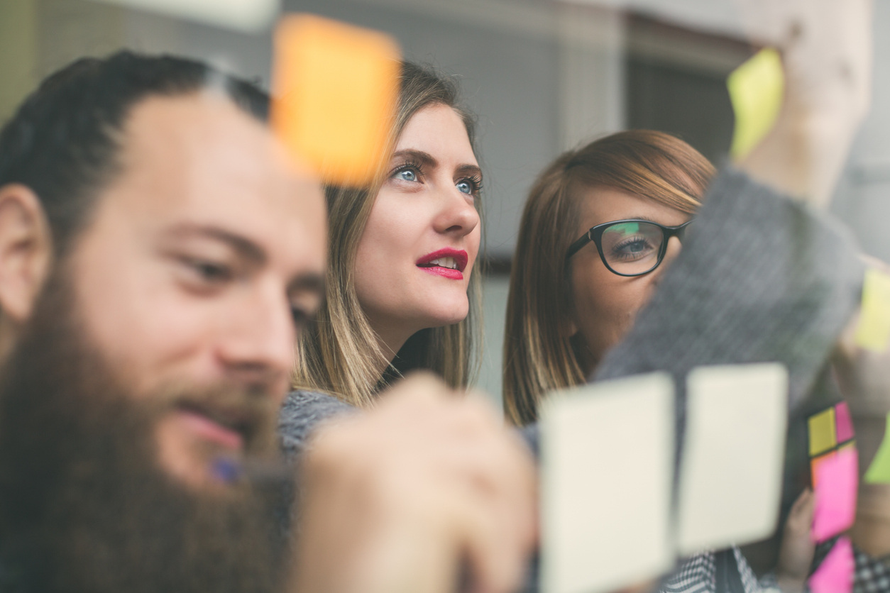 Three people, a man and two women at work all focused on post it notes. The man is writing on one the woman in the middle is looking thoughtfully at them and is also writing on one. The third only has half of her face visible as the other woman's outstretched arm obstructs it. 