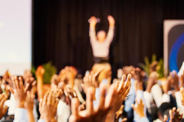 A group of raised hands with a speaker in the distance out of focus, also with raised hands. 