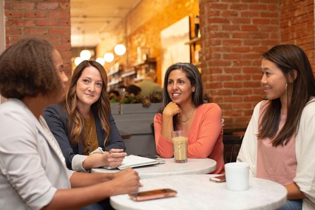 Four women at work around 2 small tables having coffee. they are all smiling and looking at one woman at the table. 