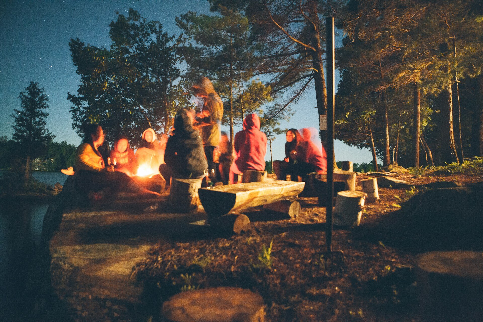 A group of people who's faces we can't see, sat around a camp fire in the woods