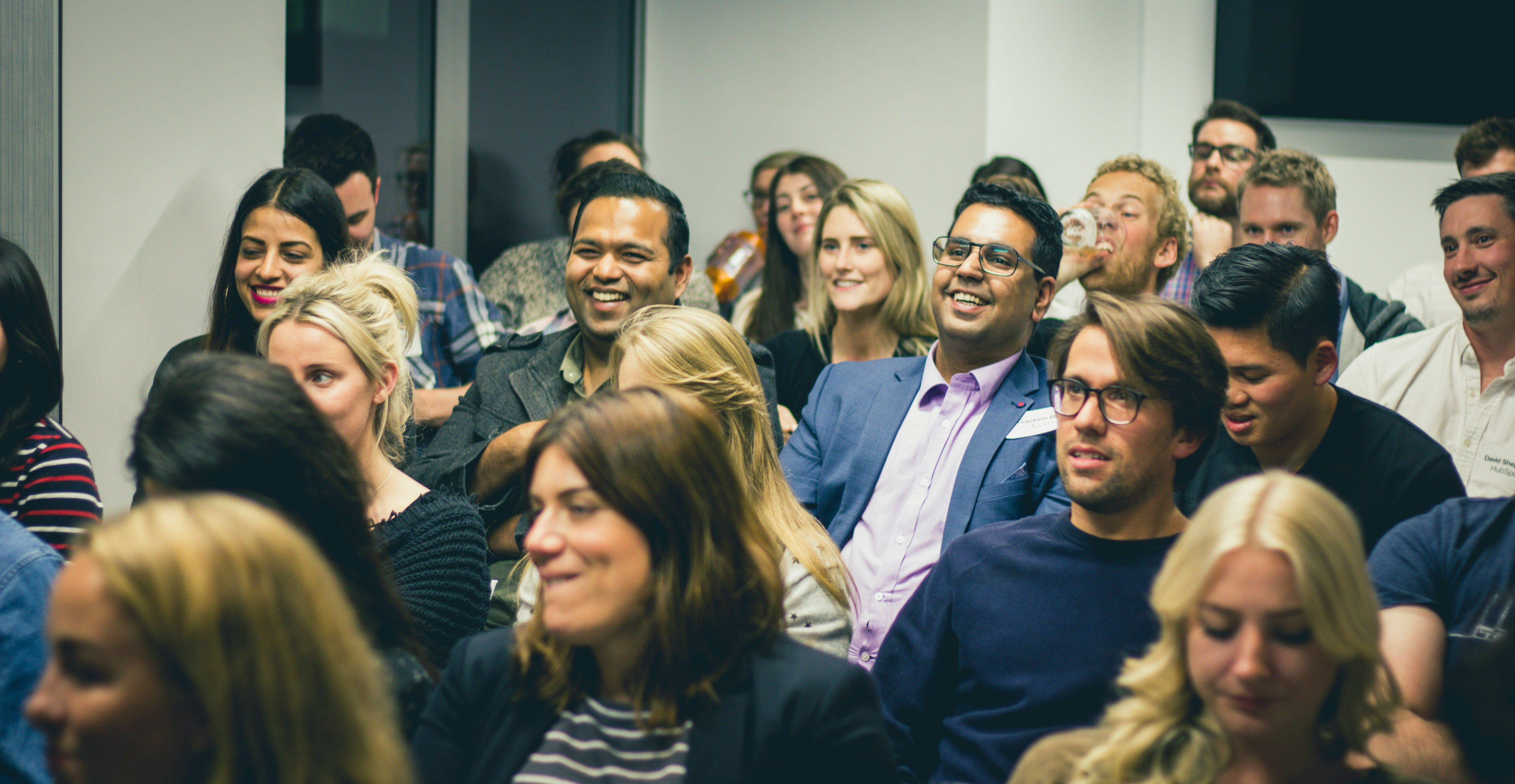 A group of people at work all sat and looking in the same direction. Some are laughing, most are smiling